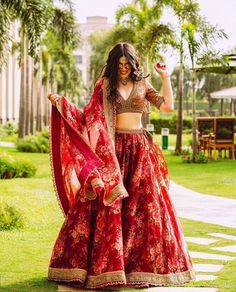 a woman in a red and gold lehenga dancing on the grass with palm trees behind her