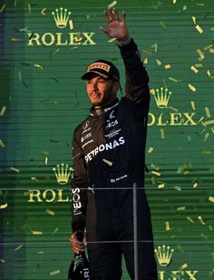 a man waves to the crowd as he stands in front of a green backdrop with gold confetti