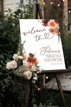 a welcome sign with flowers on it in front of some bushes and lights at an outdoor venue