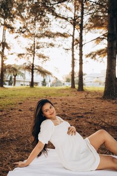 a beautiful woman laying on top of a white blanket in the grass next to trees