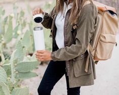 a woman with a backpack is holding a water bottle and looking at the camera while standing next to a cactus