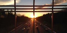 the sun is setting over an interstate with traffic going down it's sides as seen through a metal fence