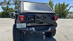 the back end of a black truck parked in a parking lot with palm trees behind it