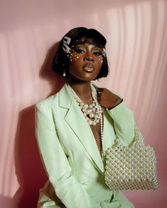 a woman in a green suit with pearls on her head and purse sitting against a pink wall