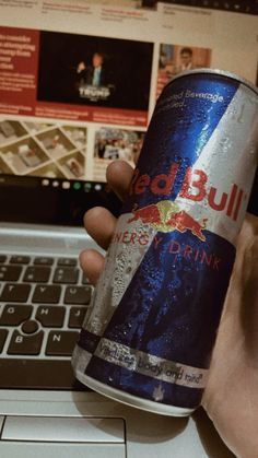 a person holding a can of red bull beer in front of a laptop