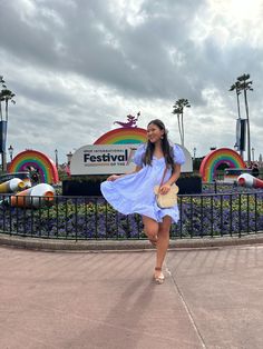 a woman in a blue dress is walking near a carnival sign and rainbow - shaped rides