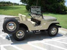 an old jeep is parked in a parking lot next to some grass and trees,