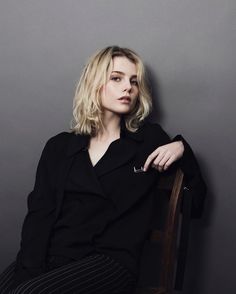 a woman sitting on top of a wooden chair in front of a gray wall with her arms crossed