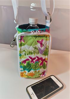 "Have you ever wished for a hands free way to carry around your water or soda bottle?   This holder is insulated to keep your water cool.  It has an adjustable strap to allow you to carry it off your shoulder or crossed over your chest.  The bottle holder has a zipper pocket for your phone or eyeglasses.  Inside the pocket there are pockets for cash or credit cards.    It also has a spring clip for your keys. This bottle holder was made from white cotton fabric with all over bright flowers.  The Water Bottle Carrier, Drinks Bottle, Soda Bottle, White Cotton Fabric, Bottle Carrier, Insulated Bags, Water Bottle Holders, Soda Bottles, Bright Flowers