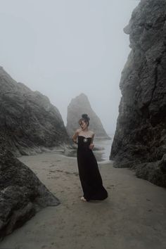 a woman in a long black dress standing on the beach next to some large rocks