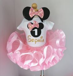 a minnie mouse birthday outfit with pink tutu and gold glitter bow on top, sitting on a wooden stand