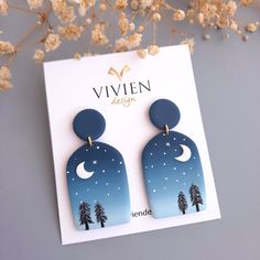 a pair of blue and white earrings with trees in the background on a card next to dried flowers