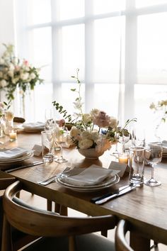 a wooden table topped with plates and vases filled with flowers next to tall windows