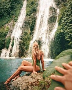 a woman in a bathing suit sitting on a rock next to a waterfall and looking at the water