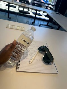 a hand holding a bottle of water next to a notebook and sunglasses on a table