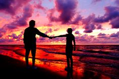 two people holding hands while standing on the beach at sunset with clouds in the background