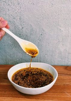 a person is spooning mustard into a white bowl on a wooden table with a concrete wall in the background