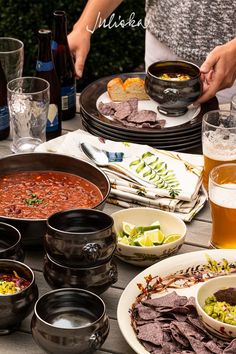 a table topped with plates and bowls filled with food next to beer glasses on top of a wooden table