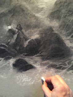 a hand holding a pencil in front of a drawing of a rocky beach and lighthouse