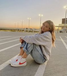 a woman sitting on the ground in a parking lot with her legs crossed and looking at the camera