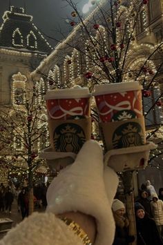 two starbucks cups are being held up in front of a building with christmas lights on it