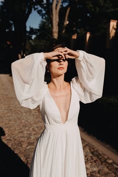 a woman in a white dress is holding her hands up to her head and looking off into the distance