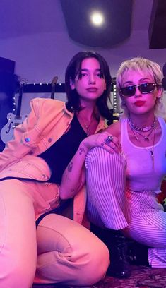 two women sitting next to each other in front of a purple light and wearing sunglasses