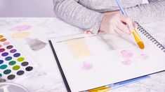 a woman is drawing on paper with colored pencils and watercolor paints while sitting at a table