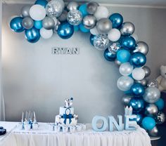 a blue and silver birthday party with balloons on the wall, white table cloths and cake