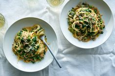 two white bowls filled with pasta and veggies on top of a table next to wine glasses