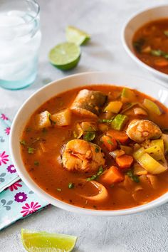 two bowls of shrimp and vegetable soup on a table with limes next to it