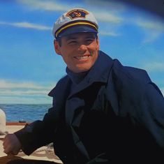 a man in a navy uniform sitting on a boat smiling at the camera while wearing a hat