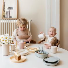 two toddlers sitting at a table eating cake and drinking milk from cups with spoons