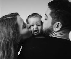 a black and white photo of a man kissing a woman's face as she kisses her baby