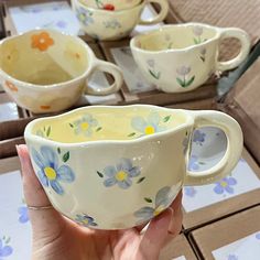 a hand holding a white cup with blue and yellow flowers painted on the side, in front of other cups