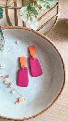 two pink and orange earrings sitting on top of a white plate next to a potted plant