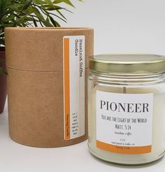 a candle next to a potted plant on a white surface with a label that says pioneers you are the light of the world