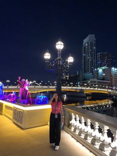 a woman standing on a bridge next to a street light and water fountain at night