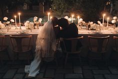 a bride and groom sitting at a dinner table with candles in the dark behind them