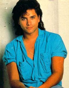 a man with long hair and blue shirt sitting on the floor in front of a white wall