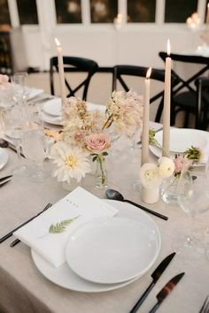 the table is set with white plates, silverware and flower centerpieces on it