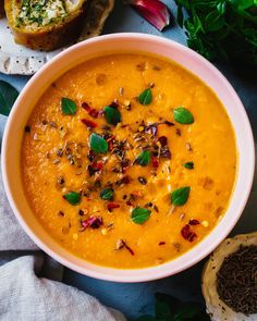 a white bowl filled with carrot soup next to some bread and green leafy greens