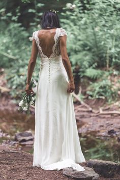 a woman in a white dress is standing on rocks by the river and looking back