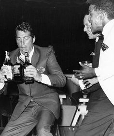 three men in tuxedos are holding beer bottles and laughing while another man sits on the floor