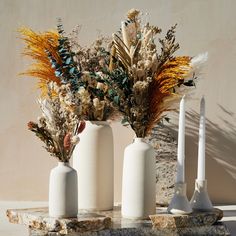 three white vases with dried flowers in them on a stone slab next to a wall