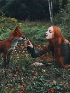 a woman with long red hair sitting on the ground next to a fox and holding it's tail