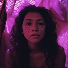 a woman with tears on her face sitting in front of a pink light and looking at the camera