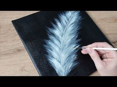 someone is painting a feather on a black canvas with white paint and a pencil in their hand