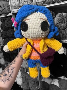 a hand holding a crocheted doll in front of a rack of yarns