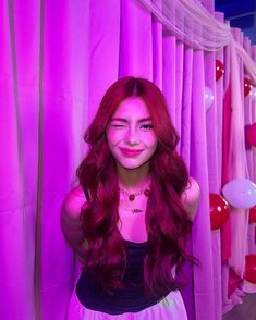 a woman with long red hair standing in front of pink curtained backdrop and balloons